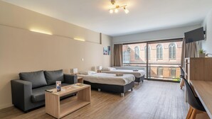 A modern hotel room with a sofa, two single beds, a desk, and a large window with a view of buildings. - Room-value Stay Residence Mechelen - Stu (Mechelen)