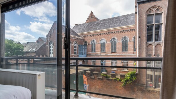 A room with a large window overlooking a historic building with arched windows and a brick facade. - Room-value Stay Residence Mechelen - Stu (Mechelen)