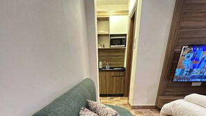 Un canapé vert avec des coussins à motifs, un téléviseur et, au fond, une petite cuisine.