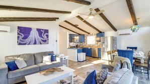 Una sala de estar moderna con un sofá gris, una mesa de centro blanca y un plaid azul estampada. Se observa una zona de cocina con armarios azules y un refrigerador de acero inoxidable.