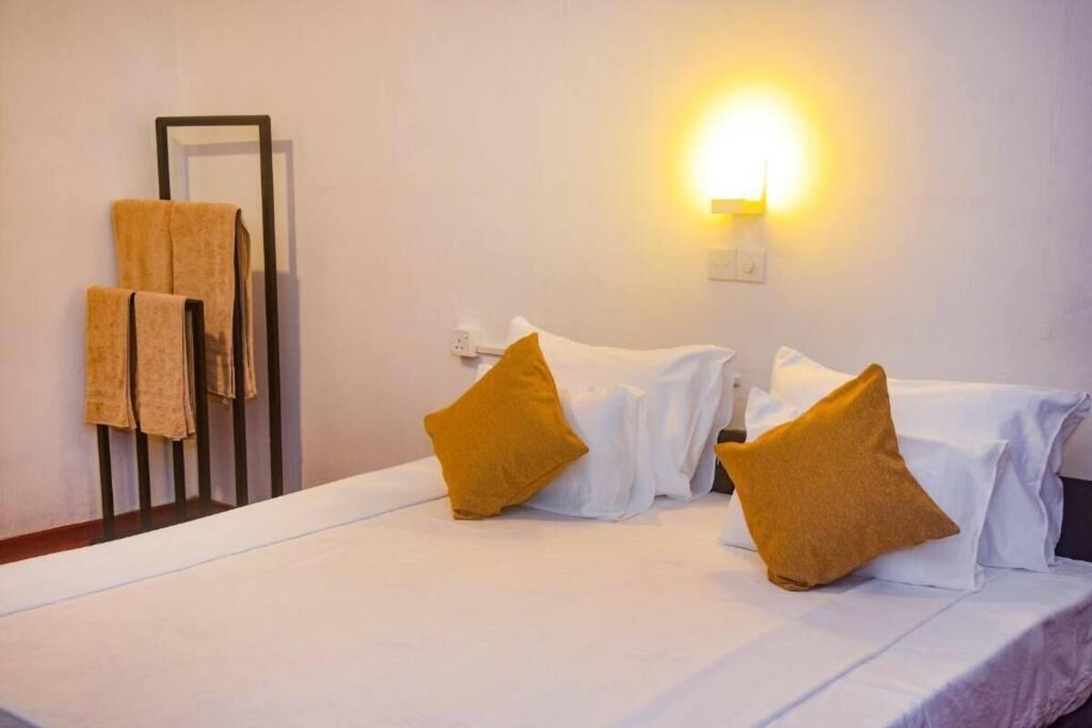 Ein Hotelzimmer mit einem Bett, weißen Laken und senfgelben Kissen. An der Wand ist eine Leuchte angebracht und es gibt einen Handtuchhalter mit Handtüchern.