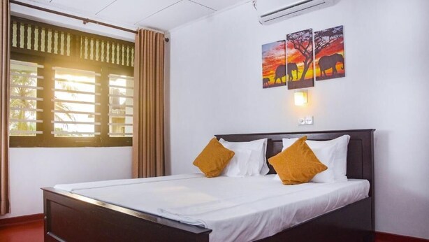 Een net opgemaakt bed met witte lakens en twee mosterdgele kussens, een raam met luxaflex en een wandairconditioning.