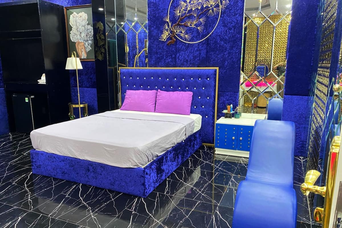 Une chambre à coucher avec un lit, une chaise bleue, une coiffeuse avec un miroir et un mur bleu orné d’un motif doré.