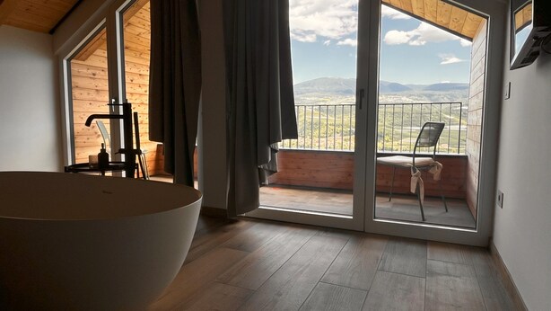 A modern bathroom with a large bathtub, wooden flooring, and a view of a balcony with a chair. - Maso Valer (Ville d'Anaunia)