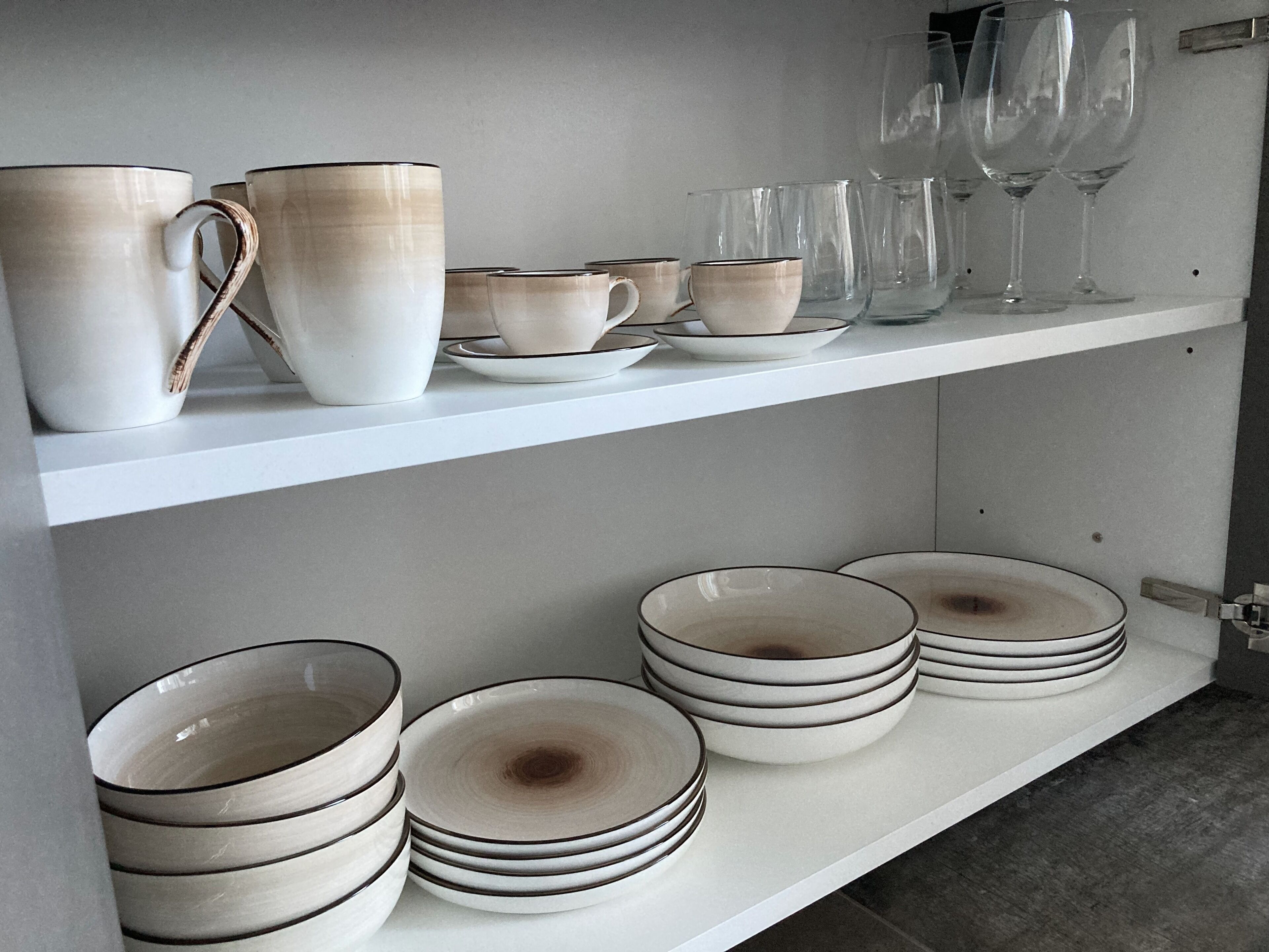 Een keukenkastje met witte en beige keramische mokken, kopjes en borden, en heldere wijnglazen.