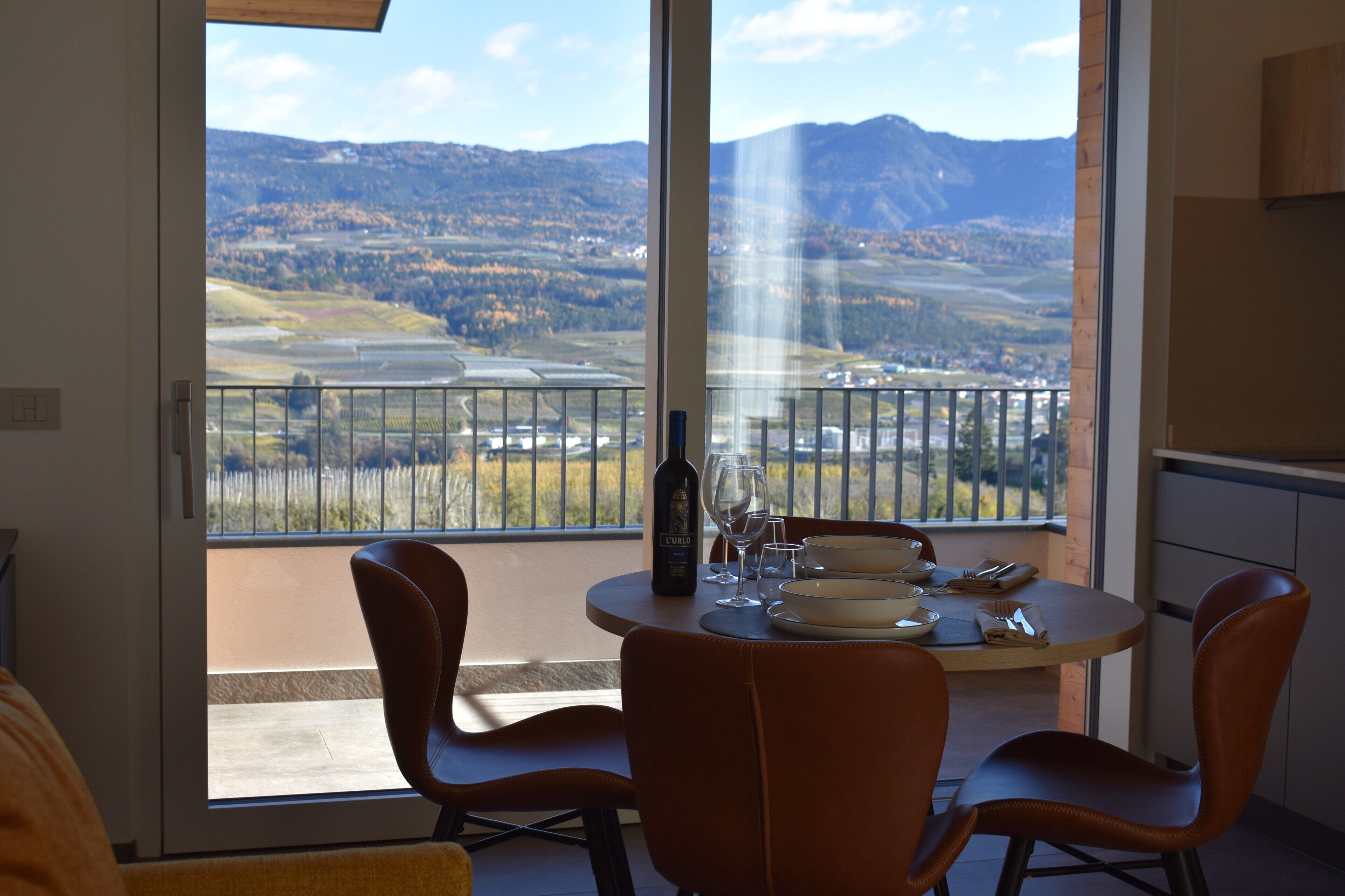 A dining area with a table set for two, a bottle of wine, and a scenic view of mountains and fields through large windows.