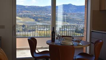 A dining area with a table set for two, a bottle of wine, and a scenic view of mountains and fields through large windows.