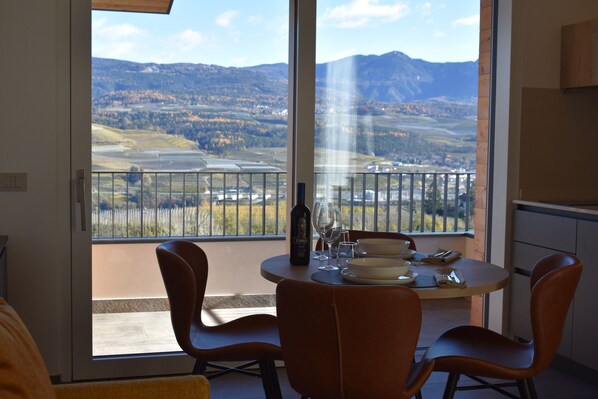A dining area with a table set for two, a bottle of wine, and a scenic view of mountains and fields through large windows. - Maso Valer (Ville d'Anaunia)