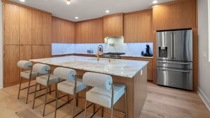 A modern kitchen with wooden cabinets, a marble island with bar stools, a stainless steel refrigerator, and a gas stove. - Contemporary Remodeled Bungalow - Pet Friendly! (San Antonio)
