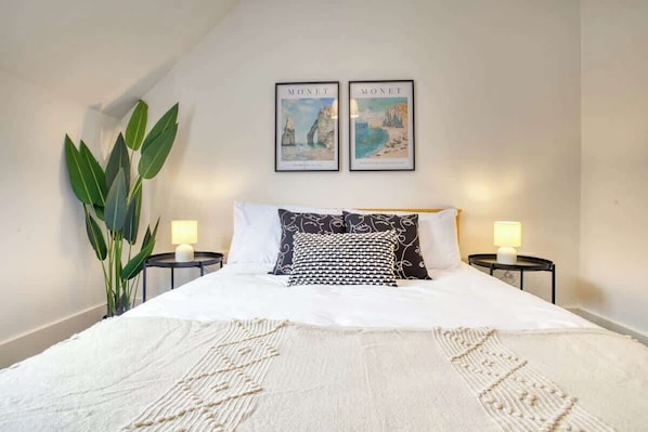 Una cama bien hecha con edredón blanco y almohadas estampadas, dos cuadros en la pared, una planta en maceta y dos lámparas de noche.