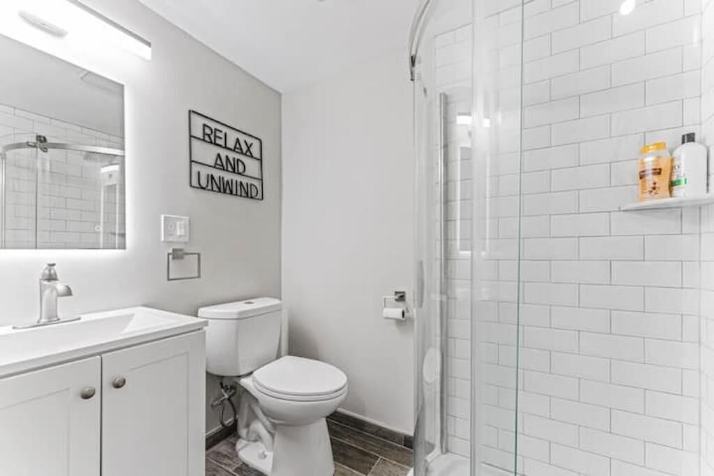Un baño moderno con lavamanos, inodoro y ducha blancos. Un cartel en la pared dice 'RELAX AND UNWIND'.
