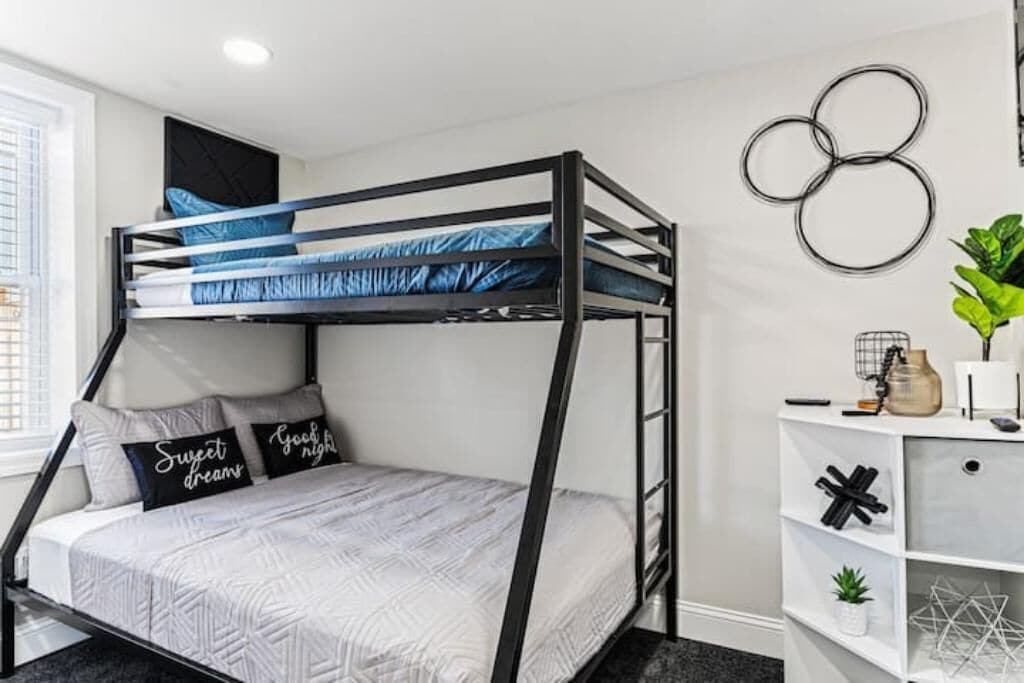 A bunk bed with a white and black frame, a patterned bedspread, and pillows with text. There is a white shelving unit with decorative items and a potted plant.
