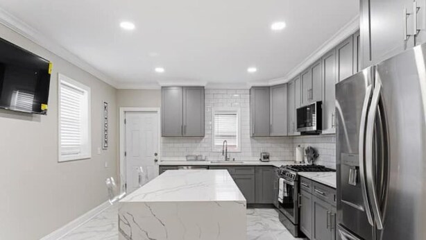 A modern kitchen with grey cabinets, a stainless steel refrigerator, a white marble island, and a tiled backsplash. - Fun, Stylish, Pool table, Darts (Camden)