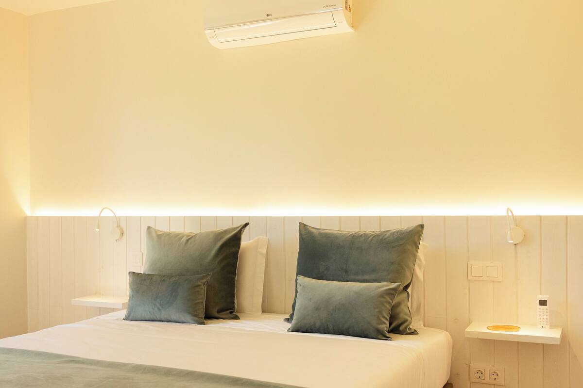 A modern hotel room with a large bed, bedside tables, a wall-mounted air conditioner, and a bedside lamp.