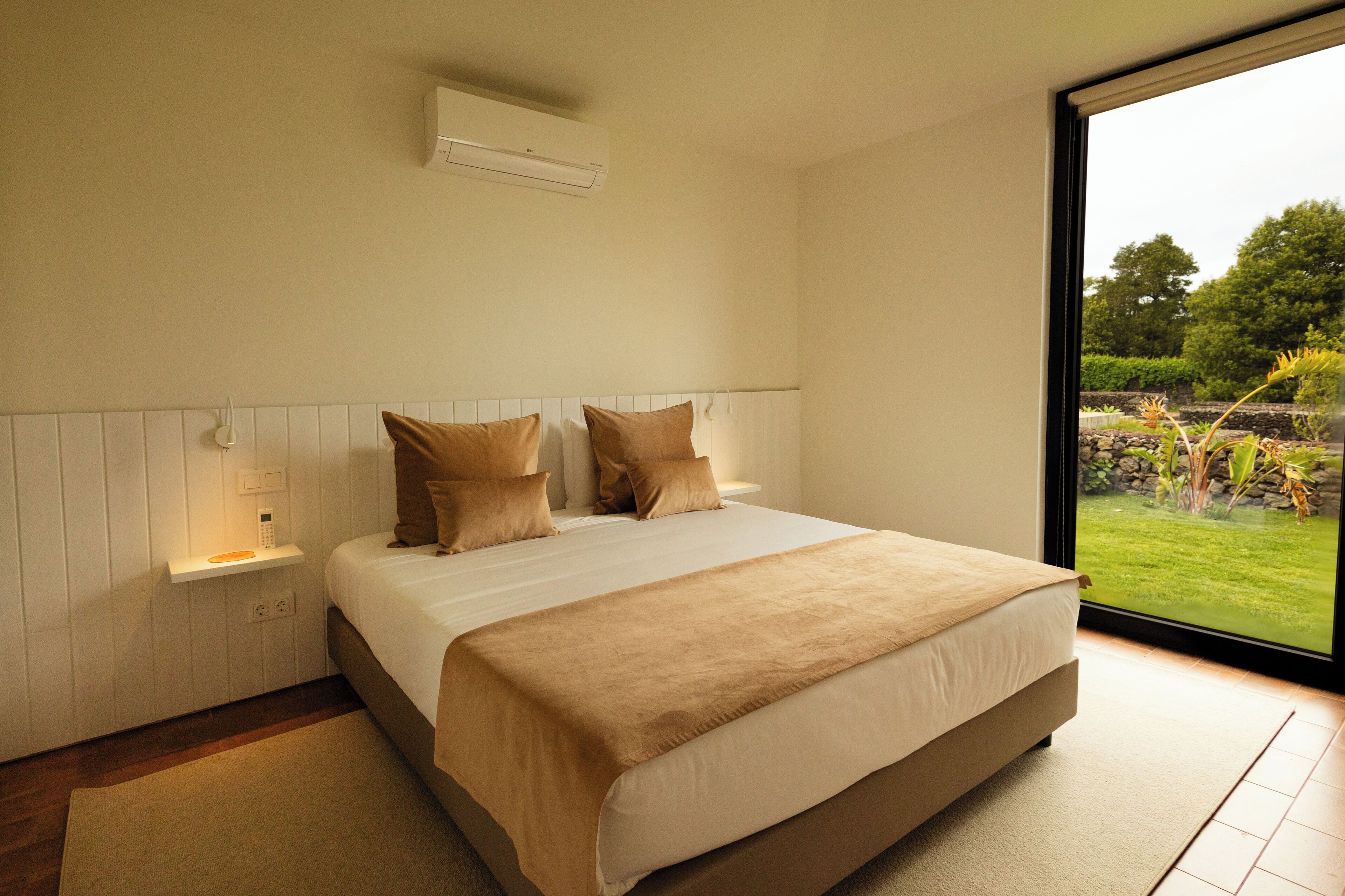 A bedroom with a large bed, a bedside table, an air conditioner, and a view of a garden through a large window.