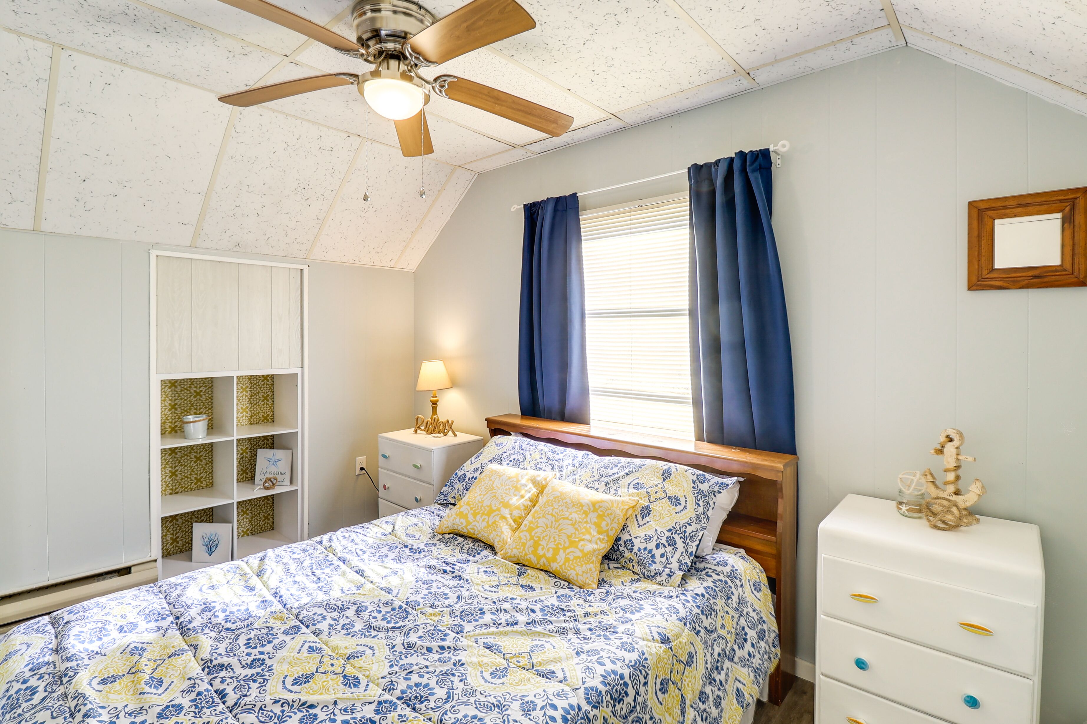 Une chambre à coucher comprenant un lit, une table de chevet, une commode, un ventilateur de plafond et une fenêtre avec des rideaux.