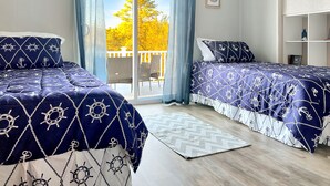 Une chambre avec deux lits, de la literie sur le thème de la marine, une grande fenêtre avec des rideaux et une vue sur les arbres.