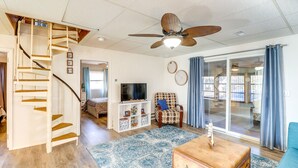 Un salon spacieux avec un escalier en bois, un ventilateur de plafond, une télévision, un tapis à motifs et un canapé bleu.