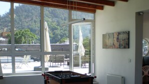 Quarto com mesa de vidro, cadeiras e mesa de futebol. Janelas amplas oferecem vista para árvores e um edifício.