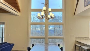 A dining area with a long wooden table, multiple chairs, a chandelier, and large windows with a view of trees. - BruinsDen- Hot Tub, Fooseball, Mountain Views (Killington)