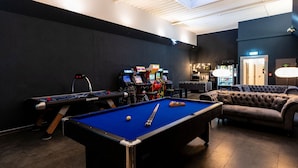 Una mesa de billar, una mesa de futbolín y varios juegos de arcade en una sala con paredes oscuras y una lámpara en el techo.