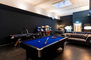 Una mesa de billar, una mesa de futbolín y varios juegos de arcade en una sala con paredes oscuras y una lámpara en el techo.