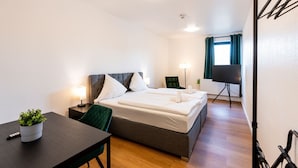 Una habitación de hotel con cama, mesita de noche, silla y televisor sobre un soporte.