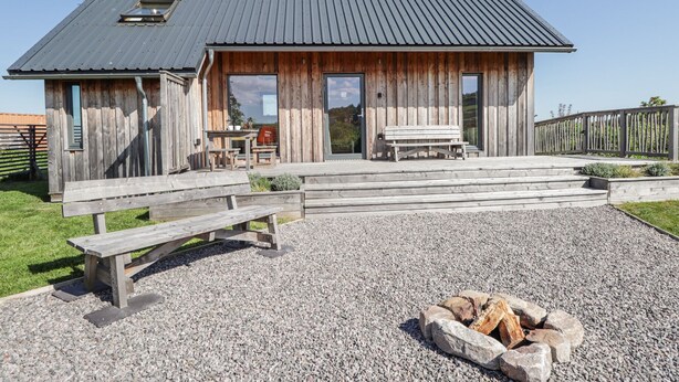 A rustic wooden cabin with a metal roof, a wooden deck, a bench, and a fire pit. - Easter Blervie (Forres)