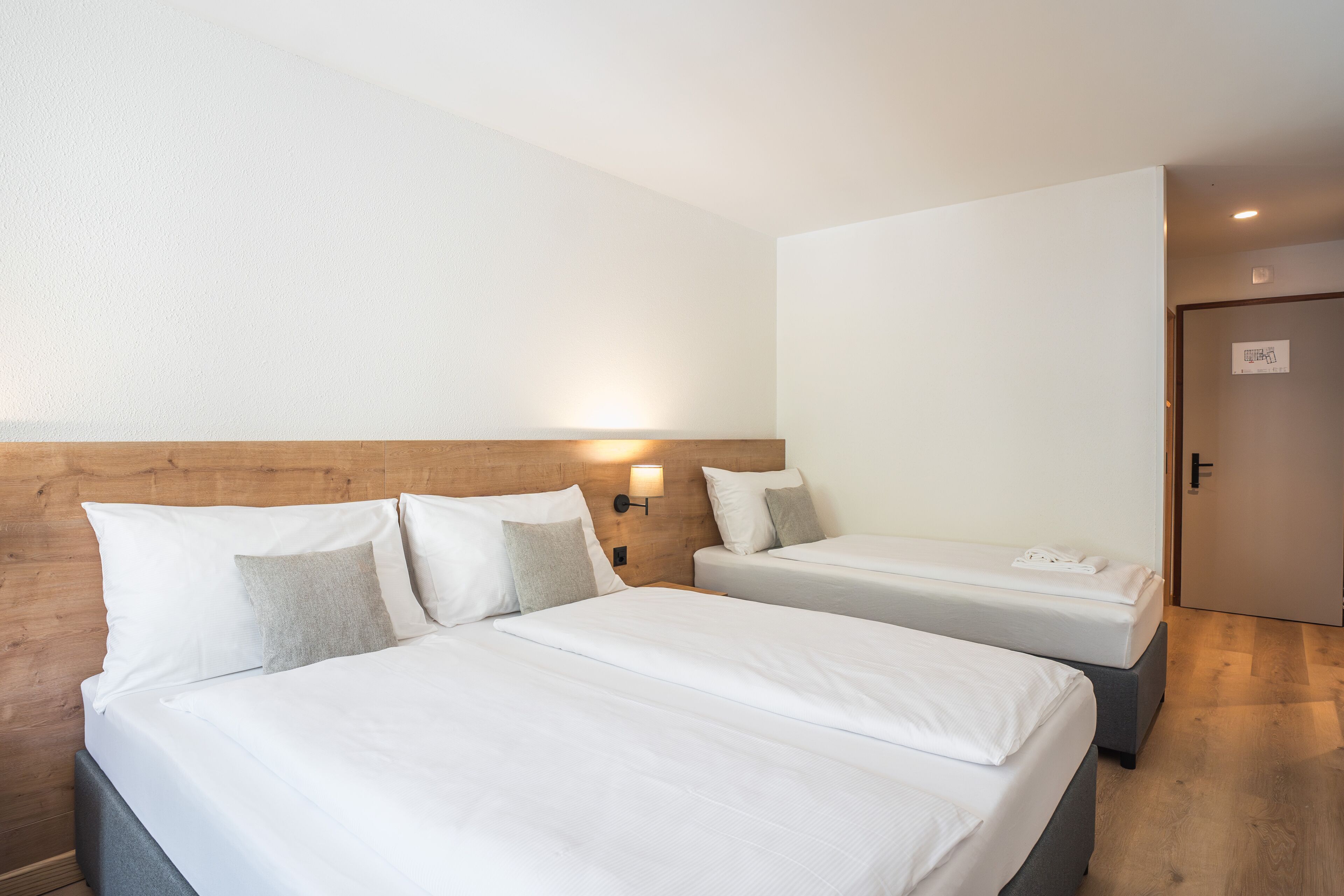 Camera d'albergo con due letti, una testiera in legno, una lampada a parete e una porta con un cartello.