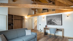 A modern living room with a grey sofa, wooden beams, a desk with a chair, and a staircase leading to an upper level.