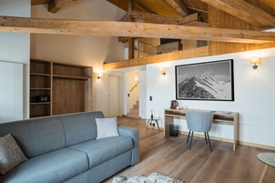 Una sala moderna con un sofá gris, vigas de madera, un escritorio con una silla y una escalera que sube a un nivel superior.