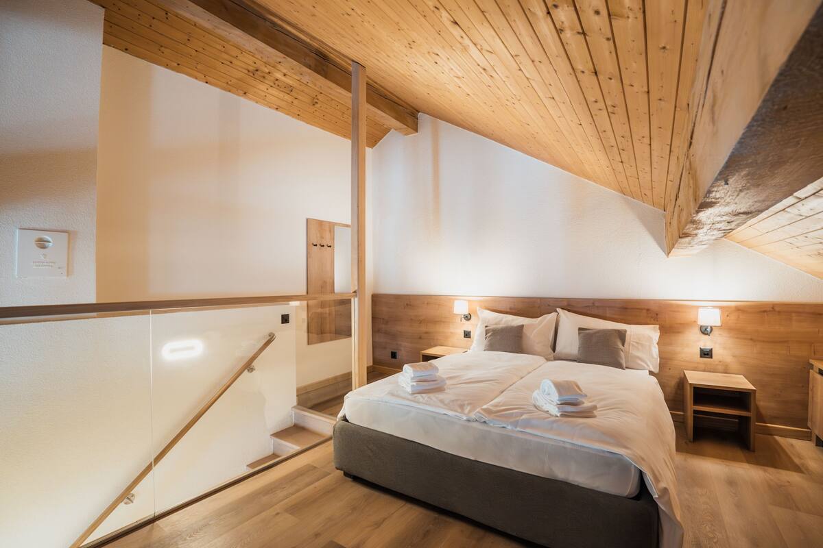 A modern bedroom with a wooden ceiling, a large bed with neatly folded towels, two bedside tables, and a wooden nightstand.