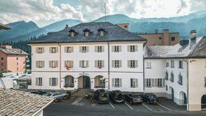 A building with a white facade, multiple windows, and a flag on top. There are parked cars in front.