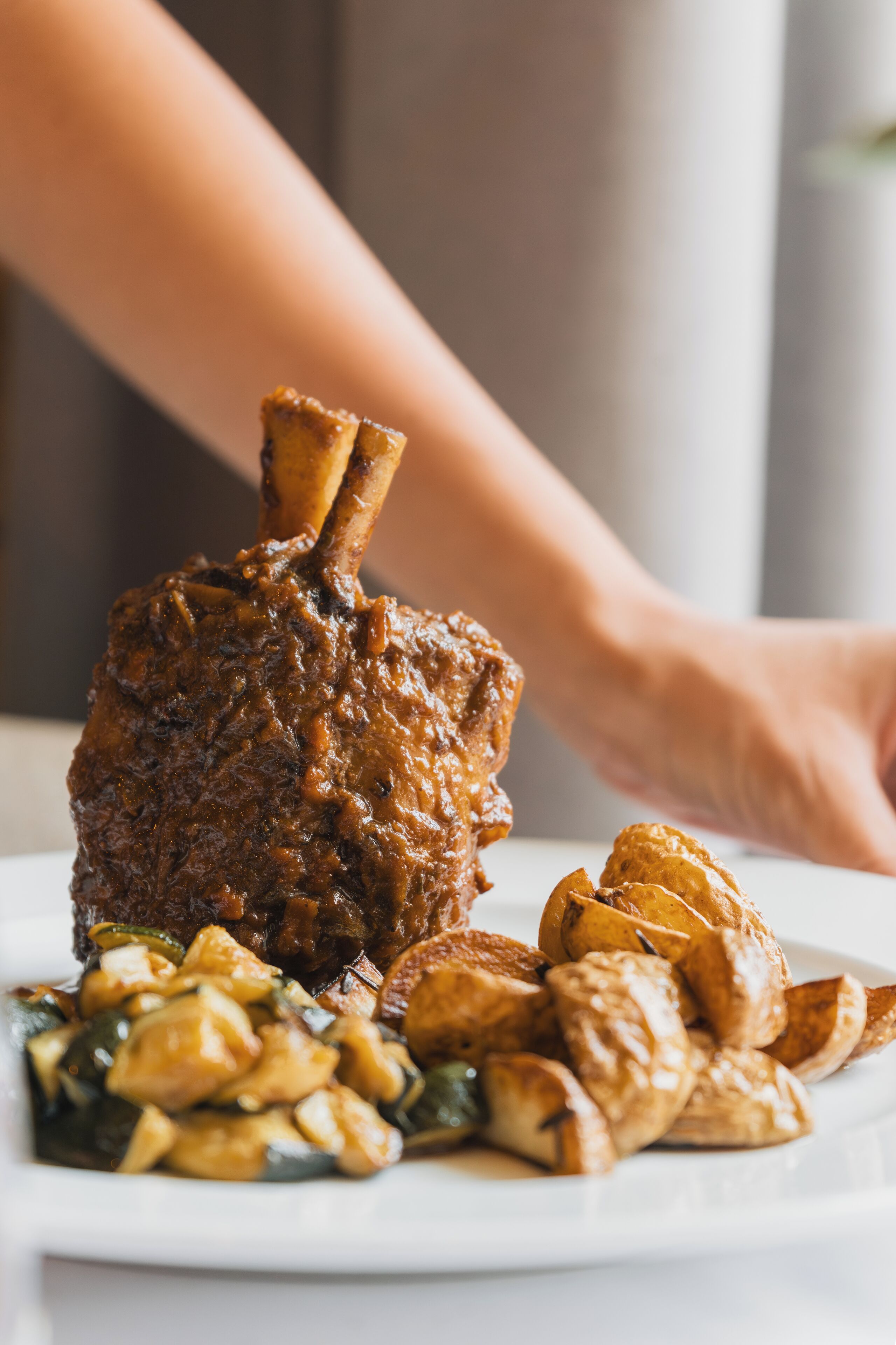A plate with a roasted meat dish, possibly a lamb shank, accompanied by roasted potatoes and a side of vegetables.