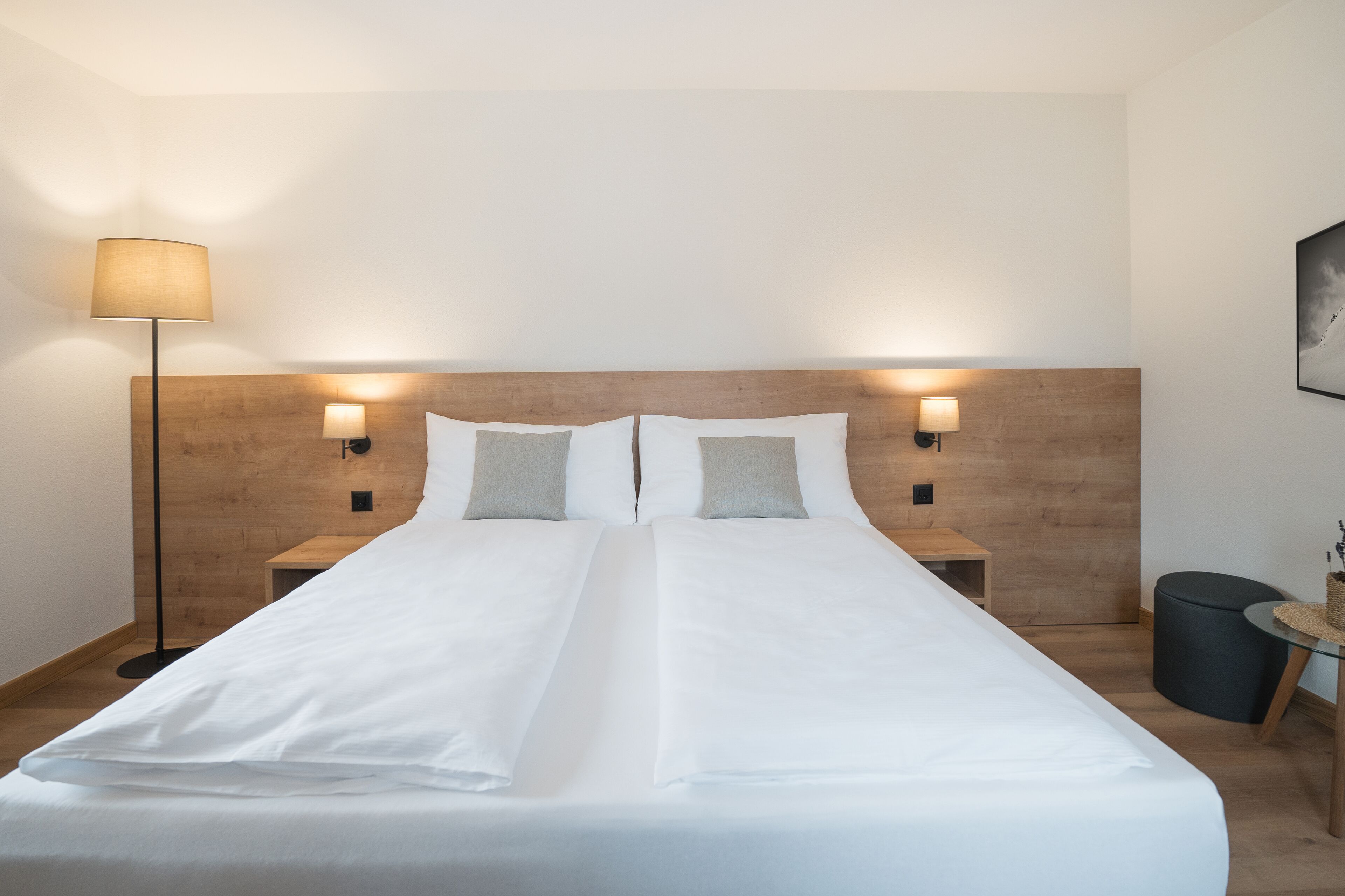 Ein ordentlich gemachtes Bett mit weißen Leinentüchern, zwei Nachttische mit Lampen, ein Holzkopfende und ein kleiner runder Tisch mit einem Korb.