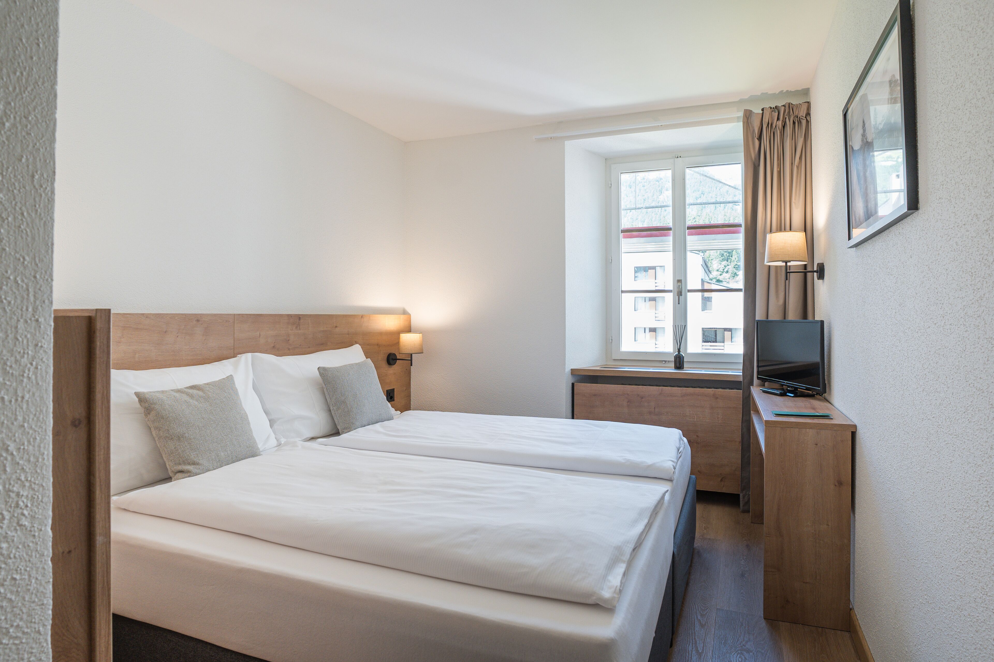 A hotel room with a double bed, a desk with a TV, and a window with curtains.