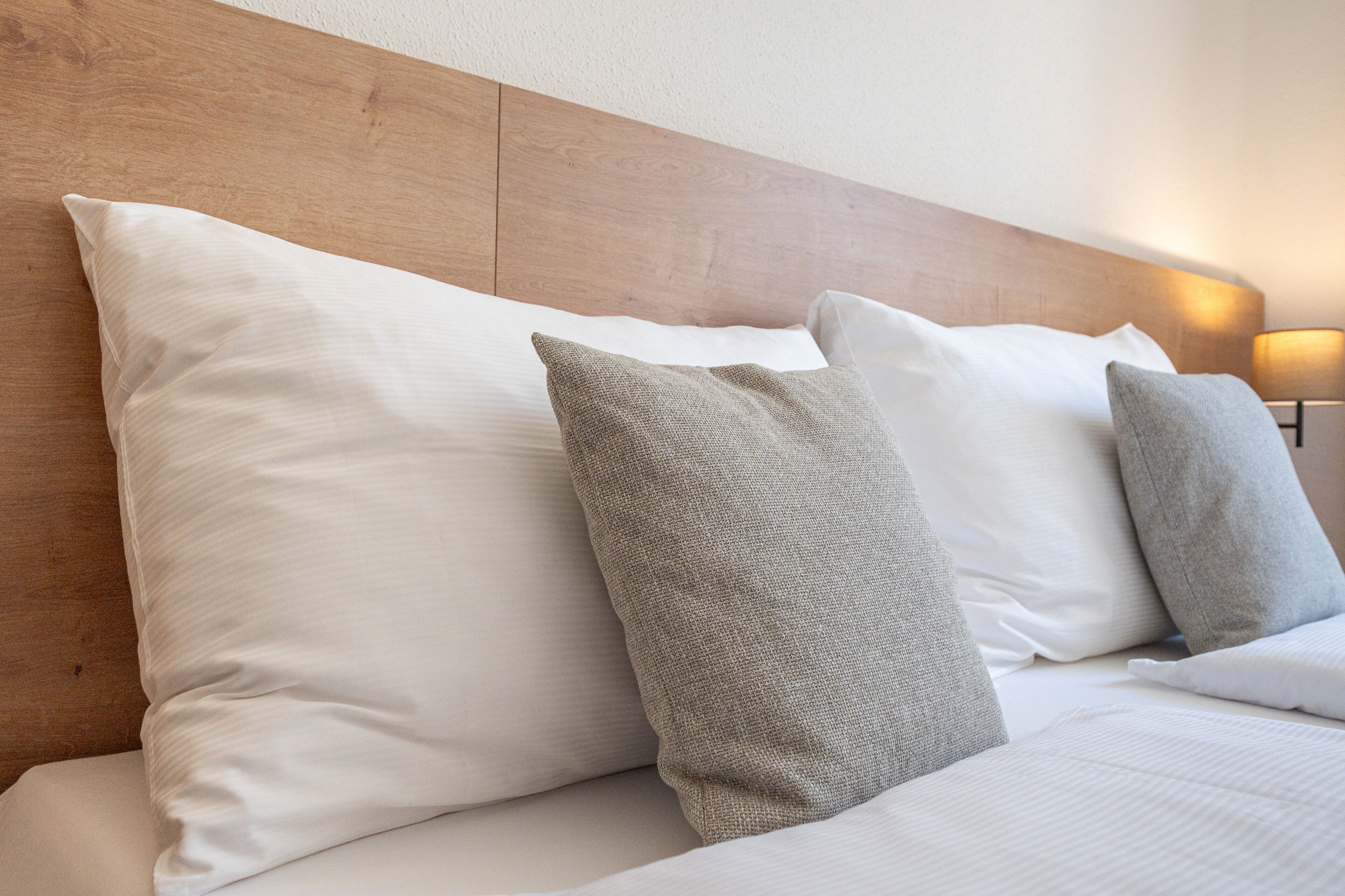 Ein ordentlich bezogenes Bett mit weißen und grauen Kissen, einem Holz-Kopfteil und einer Nachttischlampe.