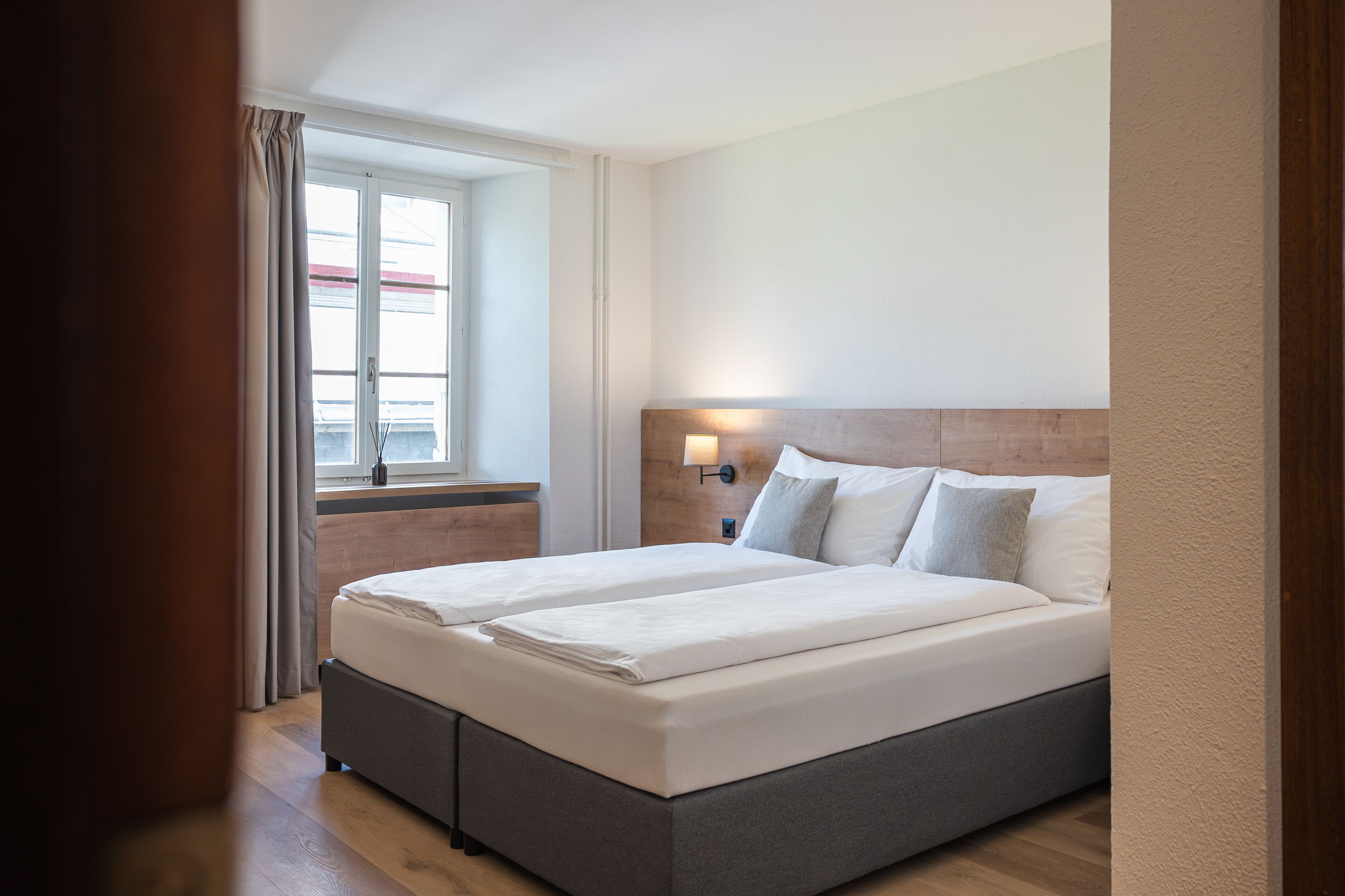 A double bed with a wooden headboard, a bedside table with a lamp, and a window with curtains.