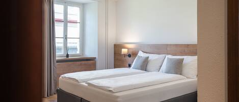 A double bed with a wooden headboard, a bedside table with a lamp, and a window with curtains.