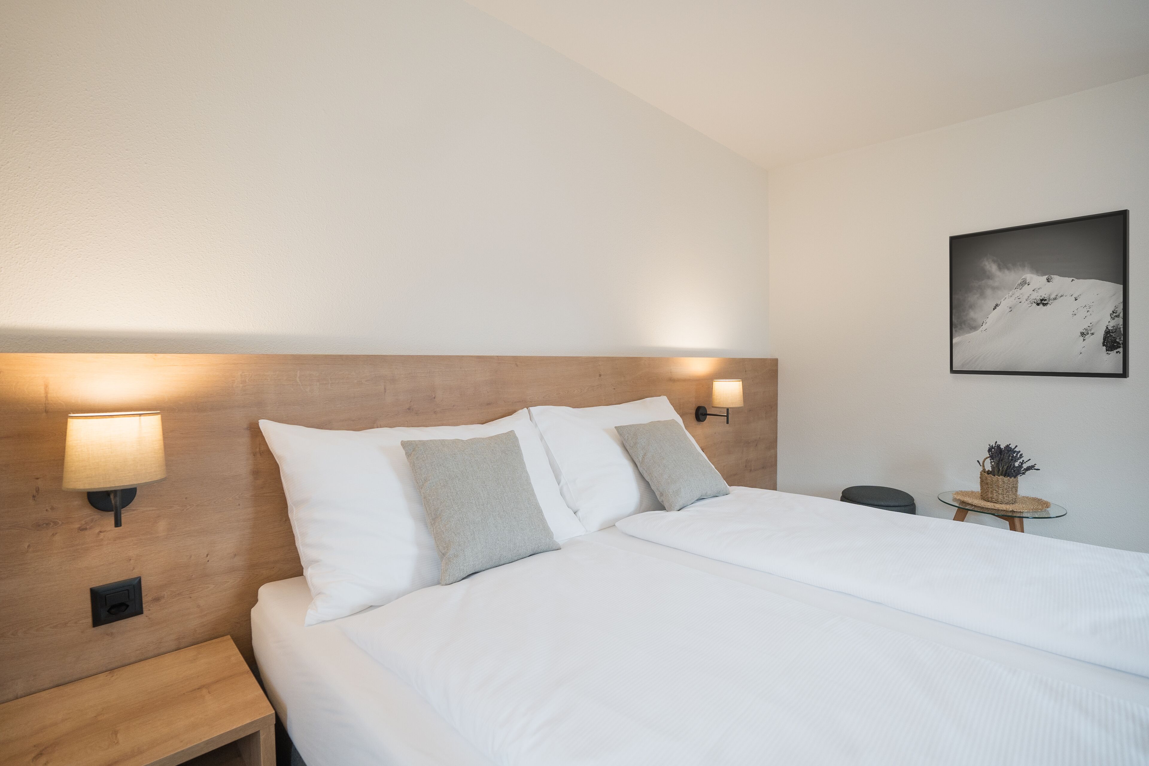 Ein ordentlich bezogenes Bett mit weißen Leinentüchern und zwei Kissen, ein hölzerner Kopfteil, eine Wandlampe und ein gerahmtes Bild an der Wand.