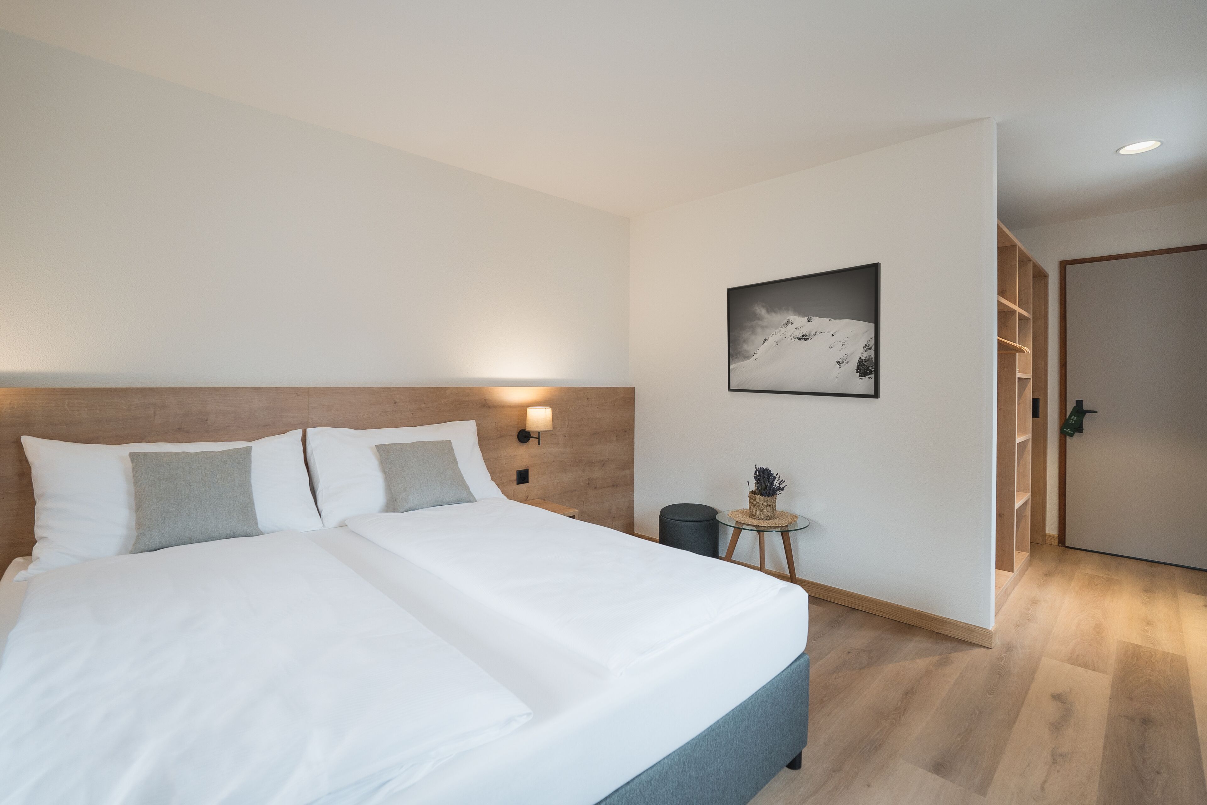 A modern hotel room with a large bed, a small side table, a wall-mounted lamp, and a framed picture on the wall.