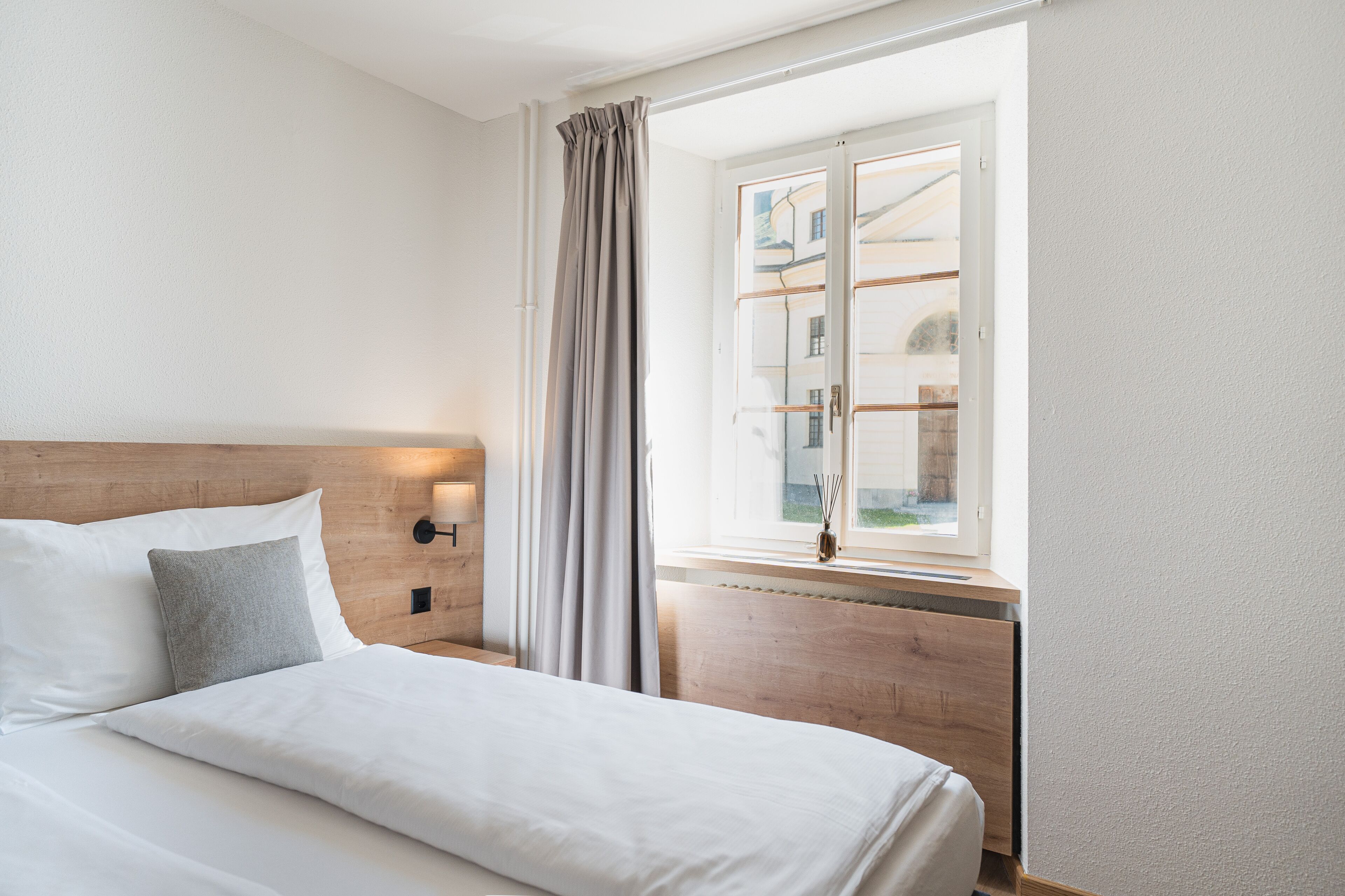 Una cama bien hecha con sábanas blancas y almohada gris, cabecera de madera, mesita de noche con lámpara, y ventana con vistas a edificios.