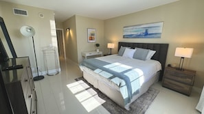 Una habitación de hotel con una cama grande, dos mesitas de noche, una lámpara de noche, un televisor montado en la pared, un ventilador de techo y un cuadro.