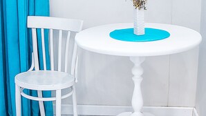 Een witte tafel met een bloevas op een blauwe mat, een witte stoel en blauwe gordijnen in een hoek.