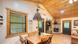 Una mesa de comedor de madera con sillas, una lámpara colgante, una chimenea de piedra y un sofá en un interior estilo cabaña.