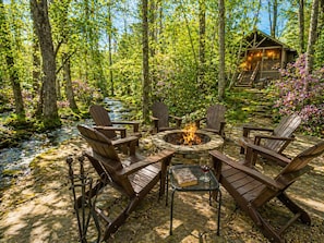 Un décor forestier avec un foyer, des chaises en bois et un chalet en arrière-plan.