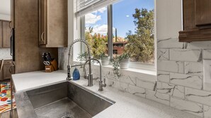 A kitchen with a stainless steel sink, wooden cabinets, and a window with a view of trees and buildings. - Andante Trail - Unit A - Red Rock Retreat (Sedona)