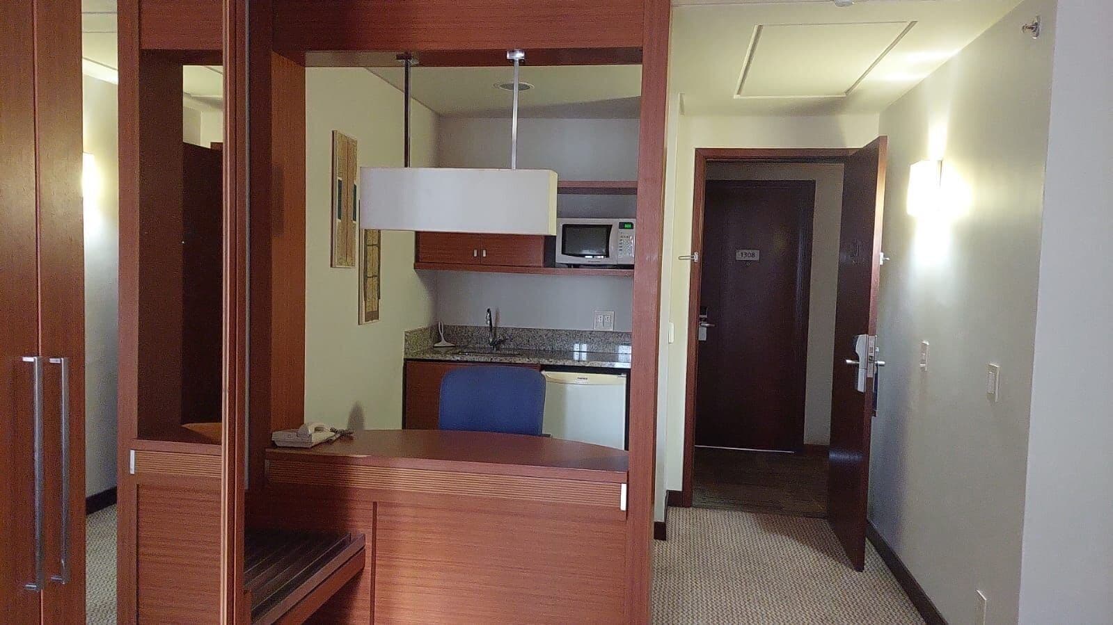 A compact hotel room with a kitchenette, a desk, and a blue chair.