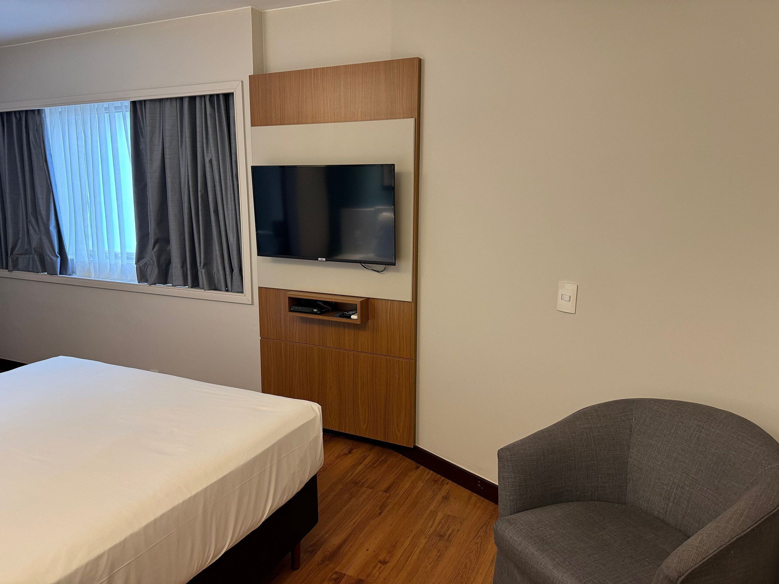 Ein Hotelzimmer mit einem Bett, einem an einer Holzplatte montierten Fernseher, einem grauen Sessel und einem Fenster mit Vorhängen.