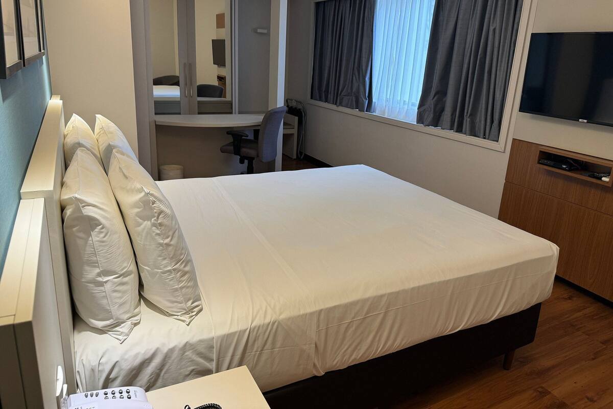 A hotel room with a large bed, a desk, a chair, a TV, and a wardrobe.